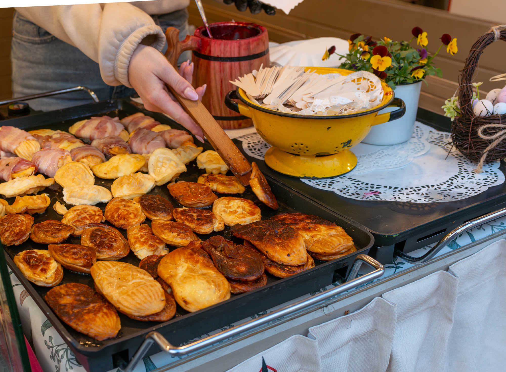 Osoba nakłada pieczone ciasteczka z dużej patelni, obok żółty garnek i dekoracyjne kwiaty